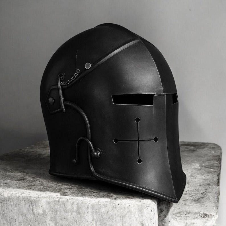 May include: A matte black metal knight helmet featuring a horizontal eye slit and a cross-shaped ventilation cutout on the front face. The helmet includes decorative metal hardware, rivets, and a small chain detail attached to the side panel.