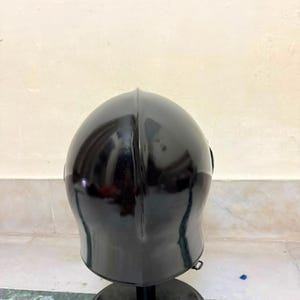 May include: A glossy black metal helmet with a smooth, rounded dome and a central vertical ridge. The helmet is mounted on a circular black stand, featuring a curved silhouette that tapers toward the base, reflecting light on its polished surface.