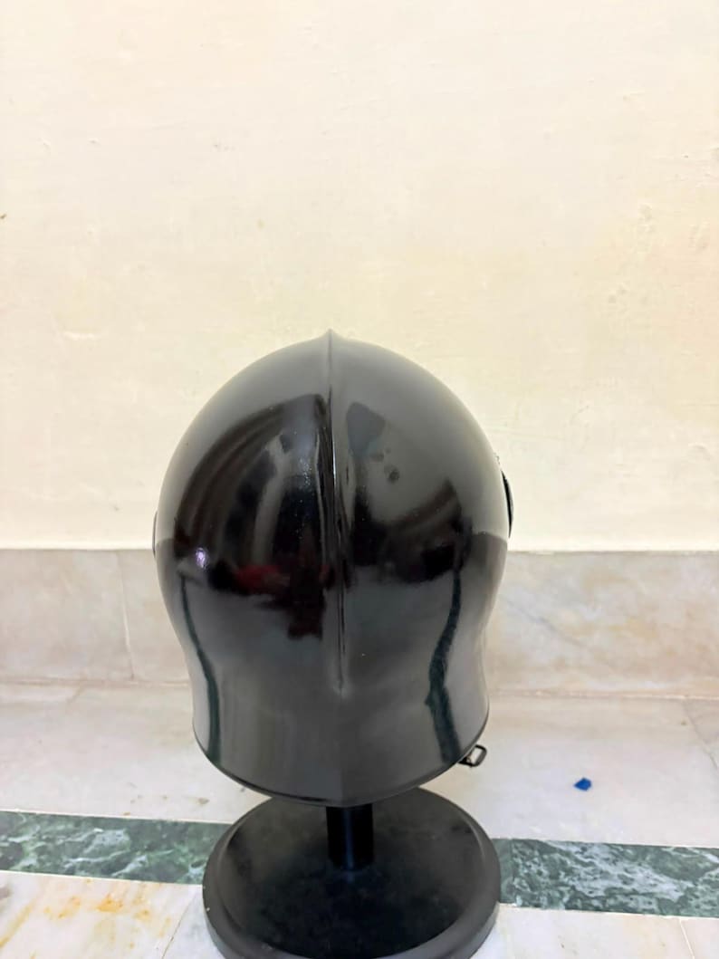 May include: A glossy black metal helmet with a smooth, rounded dome and a central vertical ridge. The helmet is mounted on a circular black stand, featuring a curved silhouette that tapers toward the base, reflecting light on its polished surface.