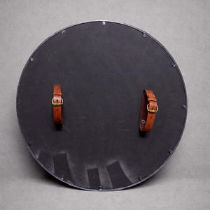 Pode incluir: Um escudo redondo preto com uma borda cinza escuro e duas correias de couro marrom com fivelas douradas. O escudo &eacute; provavelmente feito de metal e tem rebites ao redor da borda.