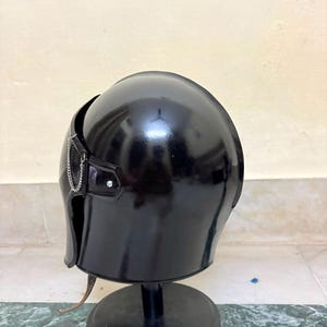 May include: A glossy black helmet with a smooth, rounded dome and a curved face guard. A thin silver-toned metal chain is attached to the side of the helmet near the temple area, secured by a small metal rivet. The helmet rests on a black stand.