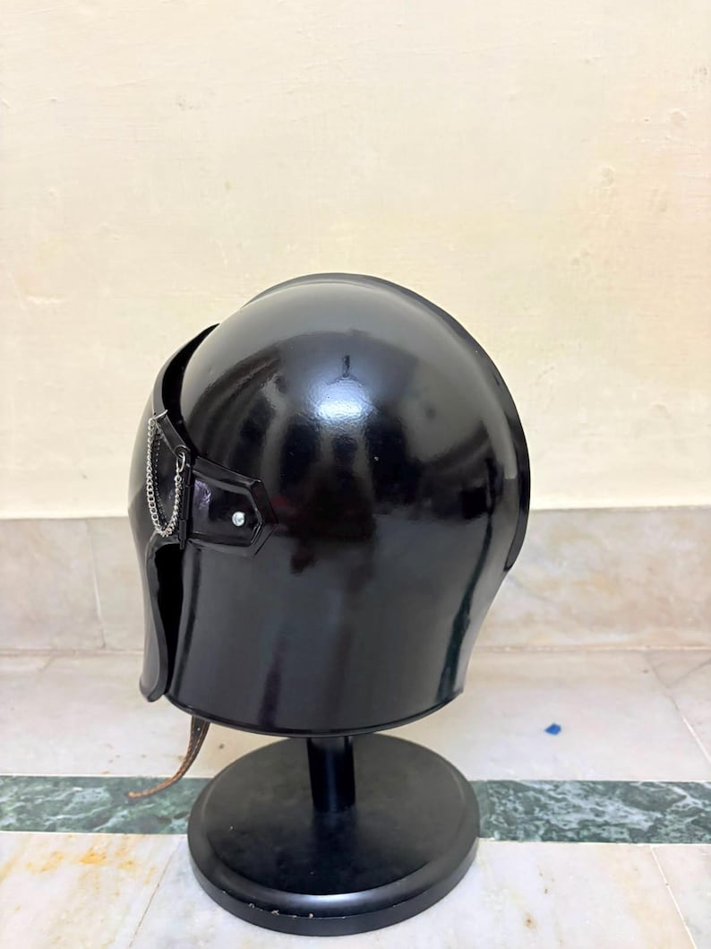May include: A glossy black helmet with a smooth, rounded dome and a curved face guard. A thin silver-toned metal chain is attached to the side of the helmet near the temple area, secured by a small metal rivet. The helmet rests on a black stand.