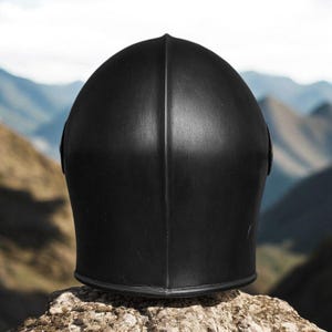 May include: A matte black helmet featuring a smooth, rounded dome shape with a prominent central vertical ridge running from the top to the base. The helmet has a solid, uniform dark finish and a curved lower edge, presenting a clean, minimalist profile.