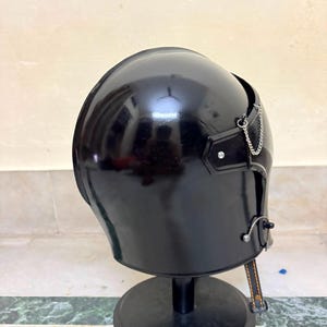 May include: A glossy black helmet with a smooth, rounded dome shape. The helmet features a small silver-toned metal chain attached to the side, a metallic fastener near the lower edge, and a thin black strap with visible orange stitching hanging down.