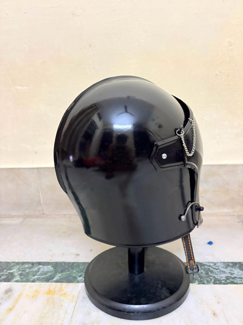 May include: A glossy black helmet with a smooth, rounded dome shape. The helmet features a small silver-toned metal chain attached to the side, a metallic fastener near the lower edge, and a thin black strap with visible orange stitching hanging down.