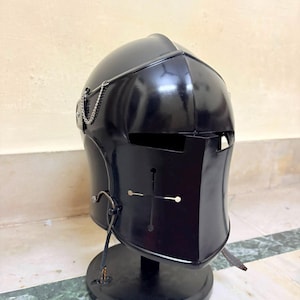 May include: A black metal knight helmet featuring a visor with a cross-shaped cutout and horizontal eye slits. The helmet includes a decorative silver-toned chain attached to the side and is mounted on a circular black display stand.