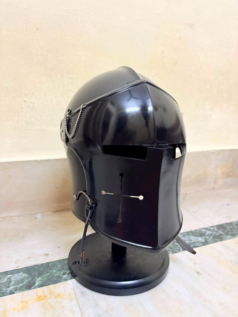 May include: A black metal knight helmet featuring a visor with a cross-shaped cutout and horizontal eye slits. The helmet includes a decorative silver-toned chain attached to the side and is mounted on a circular black display stand.