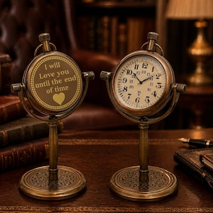 Könnte beinhalten: Zwei Schreibtischuhren im antiken Stil mit messingfarbenen Rahmen. Ein Zifferblatt zeigt ein traditionelles Zifferblattdesign, das andere den Text "I will Love you until the end of time" mit einem Herzsymbol. Beide Uhren haben verzierte Sockel.