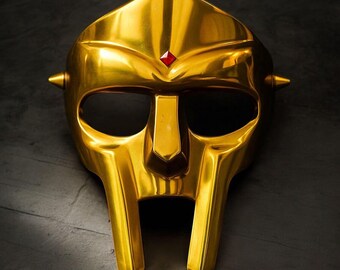 May include: A gold-colored mask with a polished finish. The mask features a pointed top, eye openings, and a prominent nose piece. A small red gem is set at the top center. Two conical spikes are on the sides.