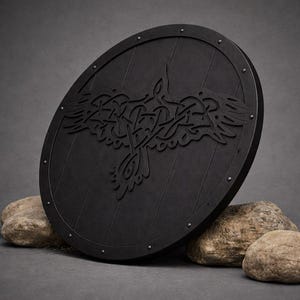Pode incluir: Um escudo redondo preto com um desenho de corvo esculpido. O escudo de madeira tem tachas de metal na borda. O corvo &eacute; retratado com intrincados n&oacute;s. O escudo est&aacute; sobre um fundo cinza com pedras.