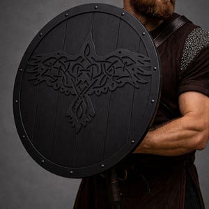 Pode incluir: Um escudo redondo de madeira preta com um desenho de corvo esculpido. O escudo tem um acabamento escuro e &eacute; adornado com tachas de metal. O corvo &eacute; retratado com intrincados n&oacute;s. Um item hist&oacute;rico ou de fantasia.
