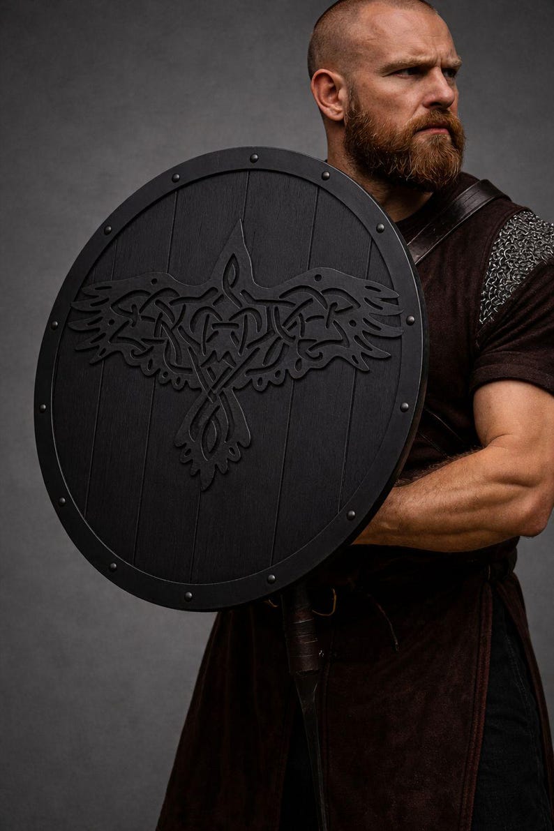 Pode incluir: Um escudo redondo de madeira preta com um desenho de corvo esculpido. O escudo tem um acabamento escuro e &eacute; adornado com tachas de metal. O corvo &eacute; retratado com intrincados n&oacute;s. Um item hist&oacute;rico ou de fantasia.
