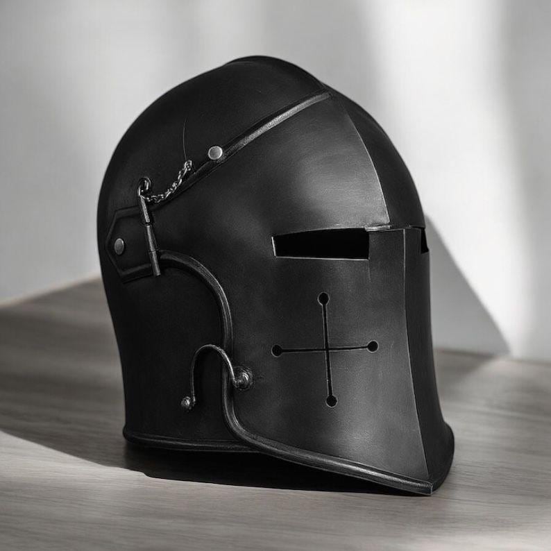 May include: A matte black knight helmet featuring a rounded dome, a horizontal eye slit, and a cross-shaped ventilation cutout on the front face. The helmet includes a small metal chain attachment on the side and silver-toned rivets.