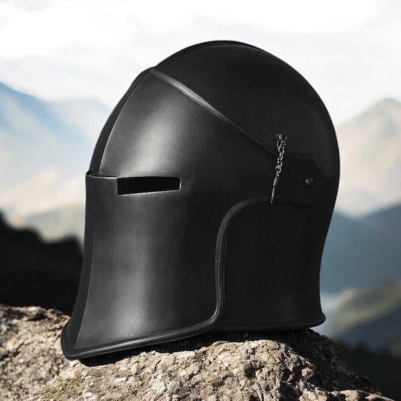 May include: A matte black knight helmet featuring a smooth, rounded dome, a horizontal rectangular visor slit, and a small metal chain attachment on the side. The helmet has a curved neck guard and a solid, uniform dark finish throughout.