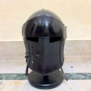May include: A black metal knight helmet featuring a smooth, rounded dome, a central ridge, and a horizontal visor slit. The front plate displays a small cross-shaped cutout with circular ends, and silver chainmail accents are visible on the sides.