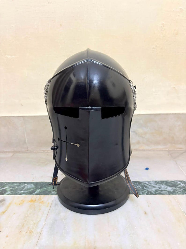 May include: A black metal knight helmet featuring a smooth, rounded dome, a central ridge, and a horizontal visor slit. The front plate displays a small cross-shaped cutout with circular ends, and silver chainmail accents are visible on the sides.