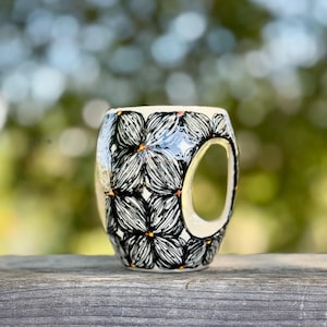 May include: A ceramic mug with a unique handle, featuring a black and white floral pattern with orange accents. The mug is cylindrical with a glossy finish, set against a blurred green and brown background.