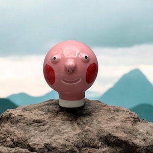 Peut inclure: Une tête en céramique rose avec un visage, des joues rouges et une base blanche. La tête est posée sur un rocher avec une chaîne de montagnes en arrière-plan.