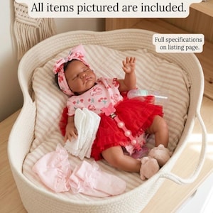 May include: A reborn baby doll in a white woven basket, wearing a pink cherry-print top, red tulle skirt, and matching headband. Accessories include a pacifier clip, a baby bottle, white cloths, pink ruffled socks, and a pink ruffled bonnet.