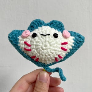 May include: Handmade crochet stingray in teal, white, pink, and red. The stingray has black eyes and a smiling mouth. The stingray is held by a hand. The stingray is a cute, handmade item.