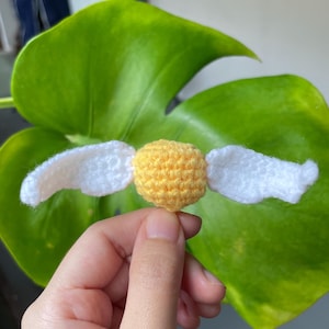 May include: A handmade, crocheted golden snitch with white wings. The snitch is a sphere with textured details, and the wings are attached on either side. The background is a large, green leaf.