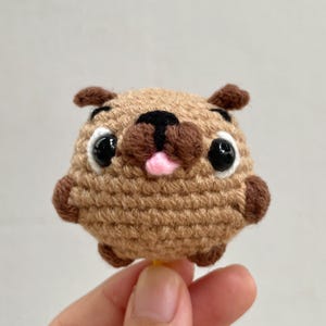 Puede incluir: Un pug hecho a mano, tejido a ganchillo, con un cuerpo marrón claro, orejas y hocico marrón oscuro y ojos grandes y negros. El pug tiene una lengua rosa y está sostenido por una mano.