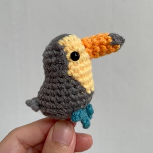 May include: A handmade crochet toucan bird with a gray body, yellow and gray head, and an orange and gray beak. The bird has a black eye and blue feet. The toucan is a small, decorative item.