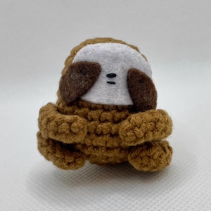 Puede incluir: Un juguete de perezoso de ganchillo hecho a mano en tonos marrón, beige y blanco. El perezoso tiene un cuerpo redondo, brazos pequeños y una cabeza con orejas marrones y una cara blanca. Los ojos están representados por dos pequeñas puntadas negras. El juguete está hecho de hilo suave.