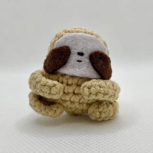 Puede incluir: Un juguete de perezoso tejido a mano en tonos beige, marrón y blanco. El perezoso tiene un cuerpo beige, orejas marrones y una cara blanca con ojos negros. El juguete está hecho de hilo suave.