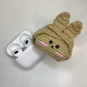 Może przedstawiać: Białe bezprzewodowe etui ładujące na słuchawki obok beżowego etui na słuchawki z szydełkowym królikiem. Etui z królikiem ma czarne oczy, różowe usta i różowe policzki. Uszy królika są wyprostowane.