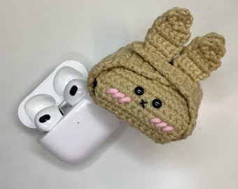 May include: A white wireless earbud charging case next to a tan crochet bunny earbud case. The bunny case has black eyes, a pink stitched mouth, and pink cheeks. The bunny ears are upright.