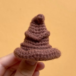 Può includere: Un piccolo cappello marrone all'uncinetto con una tesa larga e una parte superiore a punta. Il cappello è fatto di filato strutturato e sembra fatto a mano. Il cappello è una versione in miniatura di un cappello parlante, un oggetto popolare di una serie di libri fantasy.