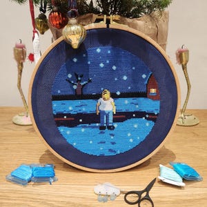 May include: A completed cross-stitch embroidery hoop with a blue background and a snowy scene. The scene depicts a lone figure standing in the snow with a house in the distance. The hoop is surrounded by embroidery floss, needles, and scissors.