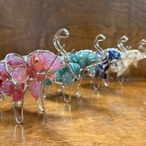 May include: A row of wire-framed elephant figurines, each filled with colorful gemstones. The elephants are in shades of pink, teal, blue, and clear, with silver wire detailing. The figurines are displayed on a wooden surface.