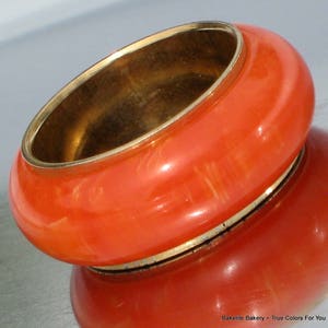 May include: A vibrant orange Bakelite bangle bracelet with a gold-tone interior and trim. The smooth, rounded design reflects light, showcasing the bracelet's glossy finish. A classic vintage accessory.