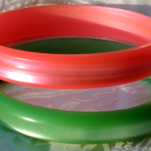 May include: Two smooth, rounded bangle bracelets stacked vertically. The top bangle is a vibrant coral red, while the bottom bangle is a deep forest green. The text "Bakelite Bakery ~ True Colors For You" is printed in the bottom right corner.