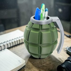 May include: A green grenade-shaped desk organizer holding pens and pencils. The organizer has a silver handle and is sitting on a wooden desk next to a notepad and other items.