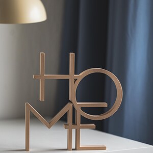 May include: A light brown, three-dimensional decorative object featuring the word HOME arranged in a stacked, interlocking block letter design. The letters are constructed with clean, straight lines and a circular O, standing upright on a flat surface.