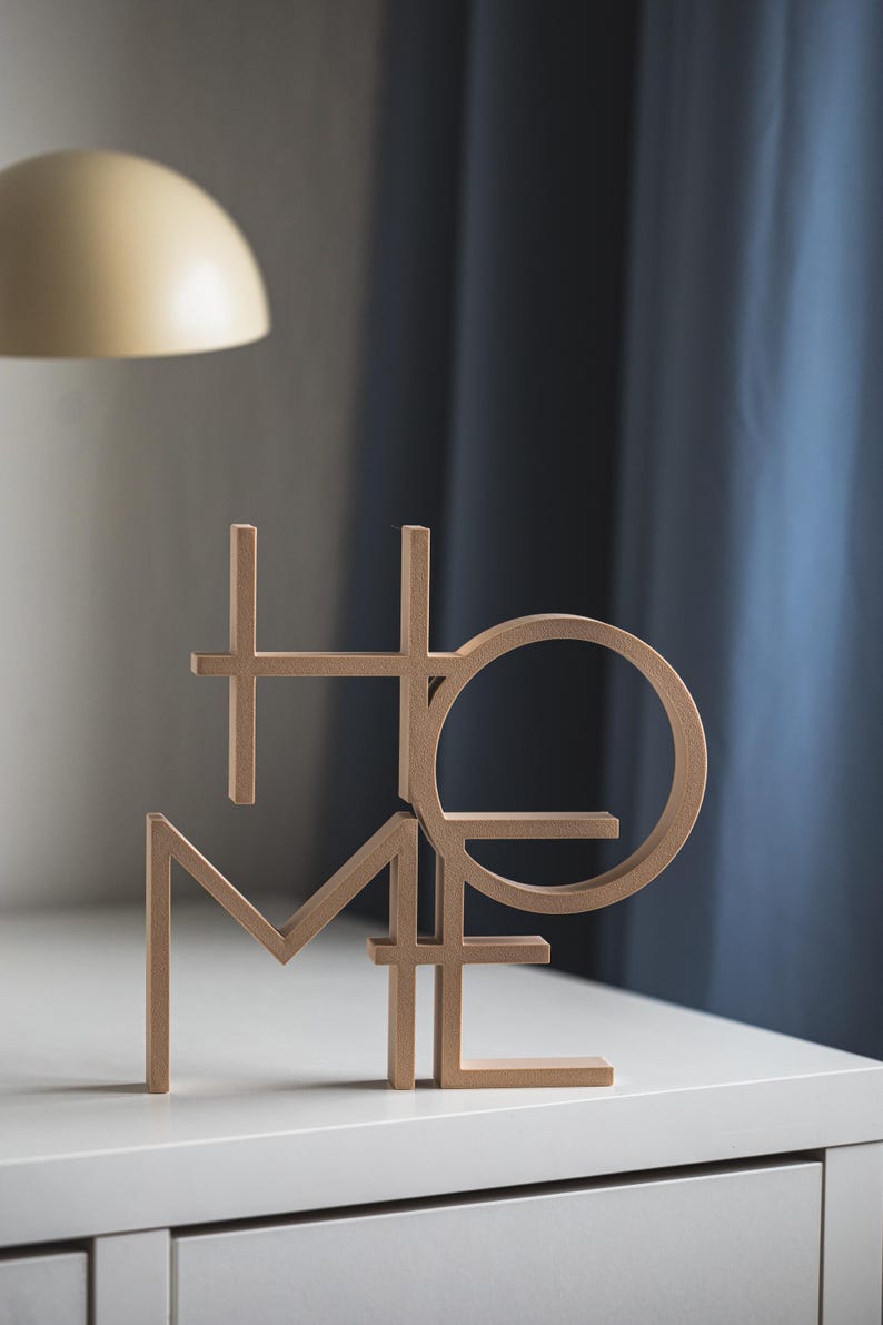 May include: A light brown, three-dimensional decorative object featuring the word HOME arranged in a stacked, interlocking block letter design. The letters are constructed with clean, straight lines and a circular O, standing upright on a flat surface.