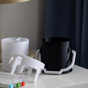 May include: Two white paint can shaped organizers with a white lid and a black paint can shaped organizer with a white lid. The organizers are on a white surface with five colorful markers.