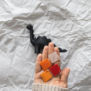 May include: A hand holding four small square samples of colored plastic. The samples are orange, red, clear, and a darker orange. The samples are on a white background.