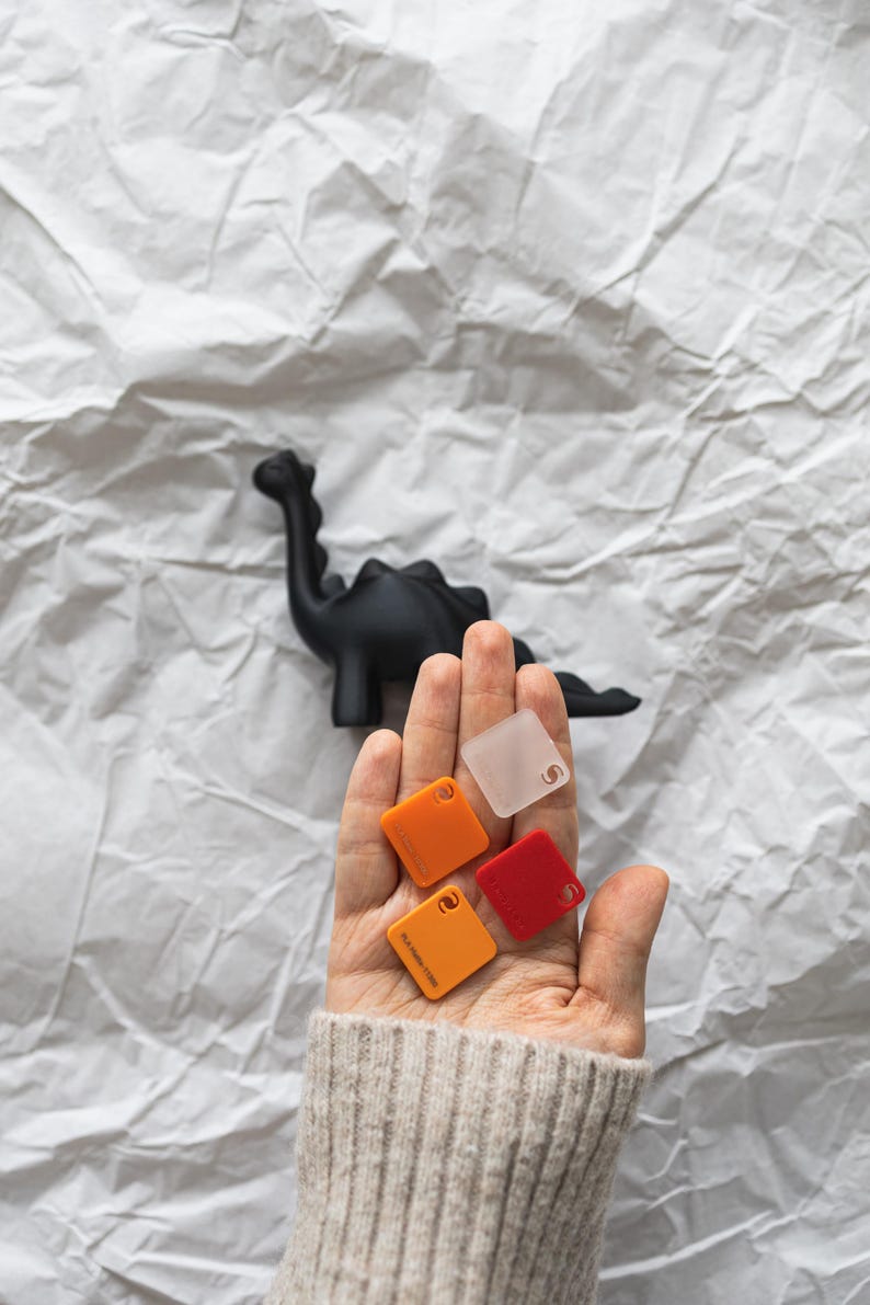 May include: A hand holding four small square samples of colored plastic. The samples are orange, red, clear, and a darker orange. The samples are on a white background.