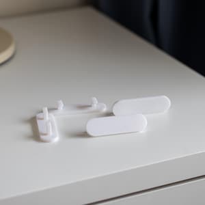 Może przedstawiać: Cztery białe plastikowe narożniki ochronne do mebli. Ochraniacze są zaprojektowane w celu zapobiegania uszkodzeniom ścian i mebli.