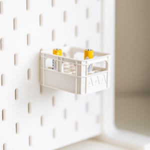 Op de afbeelding: Een witte plastic krat met twee gele AAA-batterijen erin. De krat hangt aan een wit prikbord.