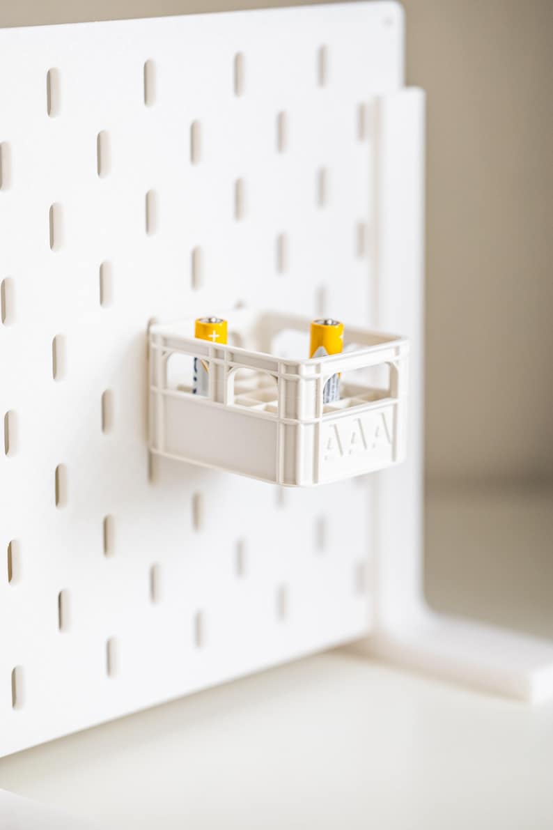 Op de afbeelding: Een witte plastic krat met twee gele AAA-batterijen erin. De krat hangt aan een wit prikbord.