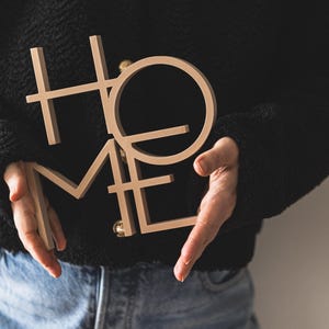 May include: A light beige, three-dimensional wooden sign featuring the word HOME in a stacked, block-letter design. The letters are connected in a single piece, with the H and O positioned above the M and E, creating a square-like composition.