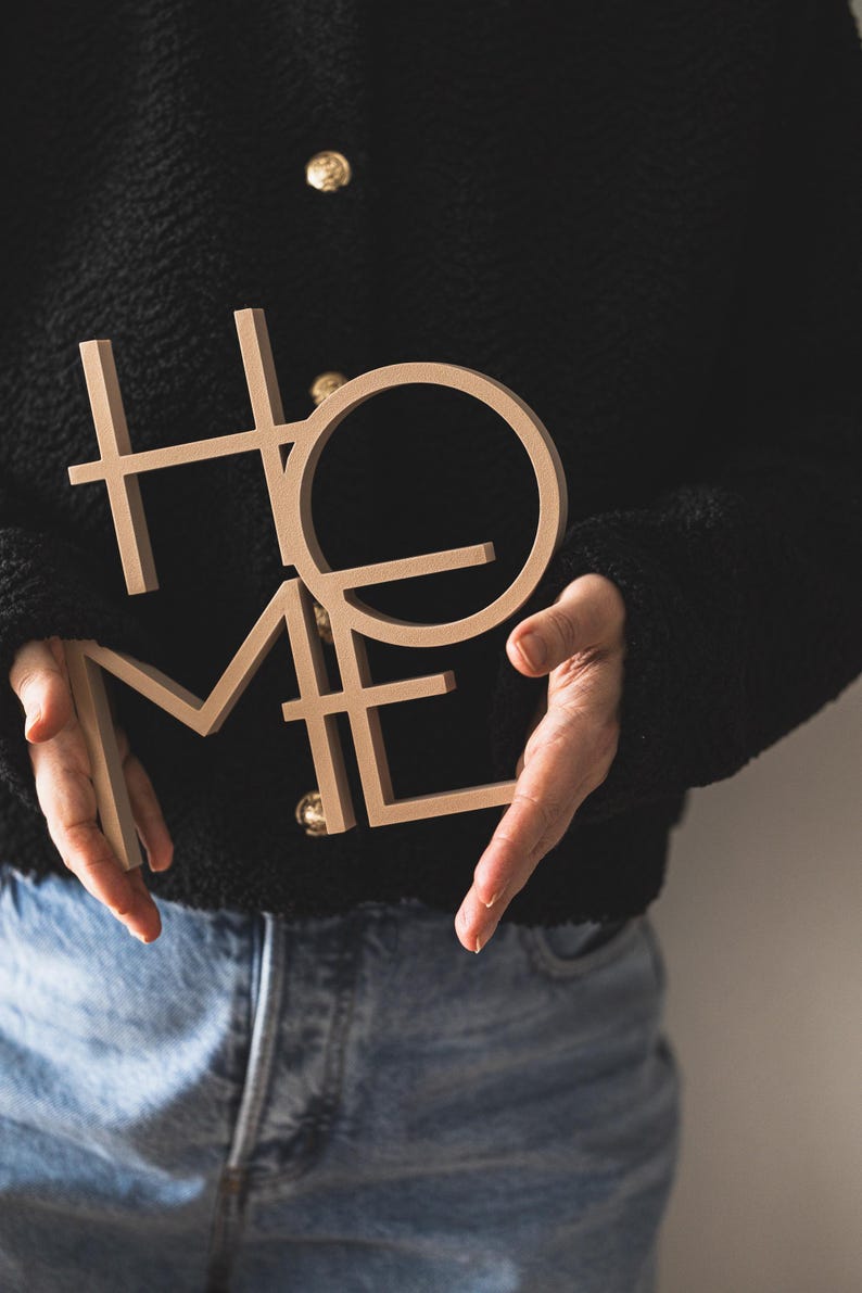 May include: A light beige, three-dimensional wooden sign featuring the word HOME in a stacked, block-letter design. The letters are connected in a single piece, with the H and O positioned above the M and E, creating a square-like composition.