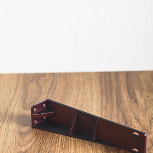 Könnte beinhalten: Eine dunkelbraune Metallhalterung mit mehreren Löchern und einem dreieckigen Design. Die Halterung liegt auf einer Holzoberfläche.