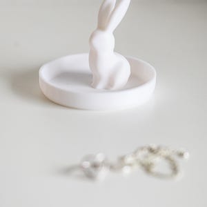 Peut inclure: Un porte-bague en c&eacute;ramique blanche avec une petite figurine de lapin blanc sur le dessus. Le plat est rond et a une surface lisse.