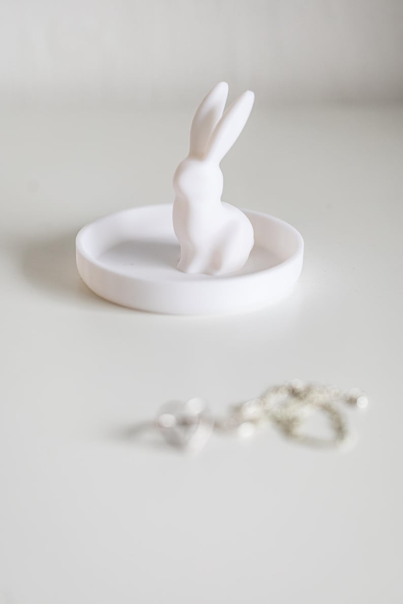 Peut inclure: Un porte-bague en c&eacute;ramique blanche avec une petite figurine de lapin blanc sur le dessus. Le plat est rond et a une surface lisse.